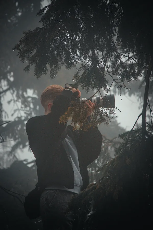 Mladý muž s fotoaparátem u oka mezi jehličnatými větvemi, v pozadí šedý les zahalený mlhou.