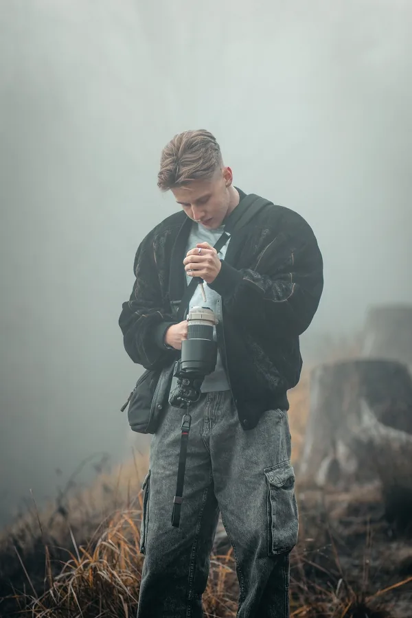 Mladý muž v tmavé bundě se dívá na fotoaparát, stojí v suché trávě, za ním hustá mlha.